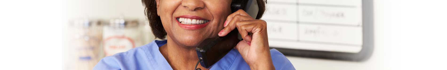 Black older nurse in blue scrubs behind computer screen holding a phone receiver for a telephone triage call