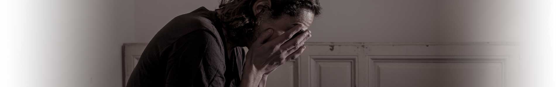 Young man in a mental health crisis sitting cross-legged on bed holding his hands in front of his face