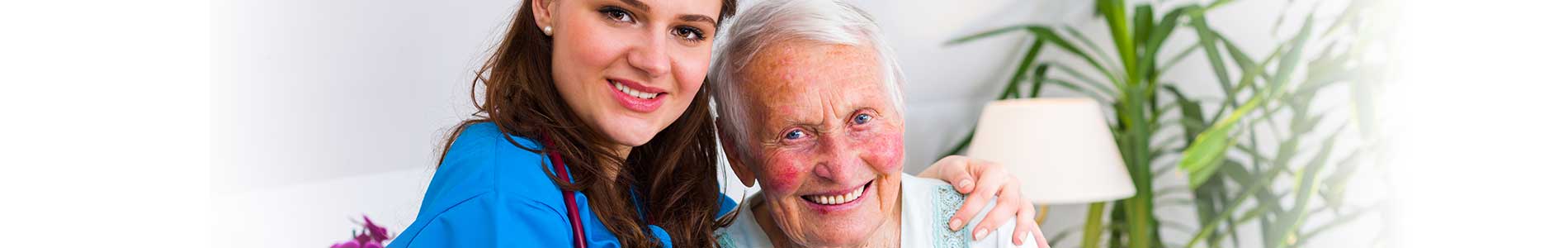 Young female nurse in blue scrubs with black stethoscope hugging and caring for elderly woman with short gray hair